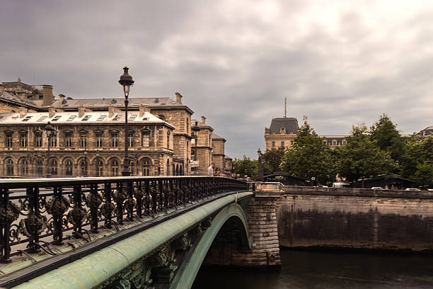 A moody daytime view of the ornate Pont au Change bridge crossing the Seine River, framed by historic stone buildings in central Paris, France. This classic Parisian scene captures French architectural elegance and urban heritage under overcast skies. A subtle blend of intricate ironwork, 19th-century facades, and atmospheric weather evokes the timeless character of the French capital.