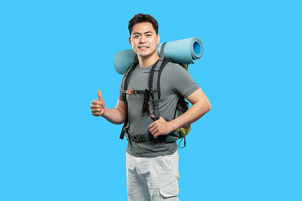 istock Smiling Asian man with backpack ready for adventure giving a thumbs up on blue background 2220743050