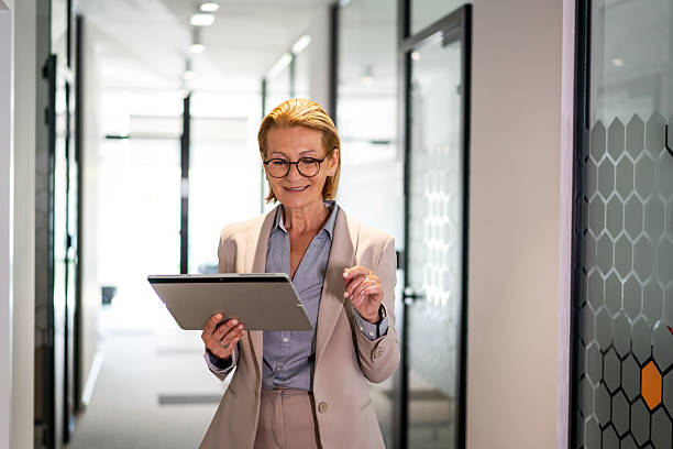 istock Smiling mature businesswoman walking through office corridor using digital tablet 2220743290