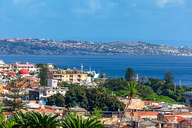 Picturesque view of Messina, Sicily, with colorful buildings along coastline and vibrant sea stretching toward horizon. In background, rolling hills dotted with homes enhance charming coastal town