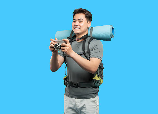 istock Smiling hiker with backpack and rolled mat uses a camera on blue backdrop capturing adventure 2220746840