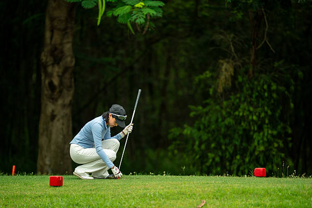 A woman is kneeling down on a golf course. She is holding a golf club and looking at the ground