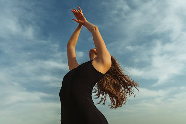 A woman wearing a black dress moves gracefully, her arms raised, framed against a vibrant blue sky. Her hair flows naturally, enhancing the dynamic and artistic composition, exuding a feeling of freedom and joy.