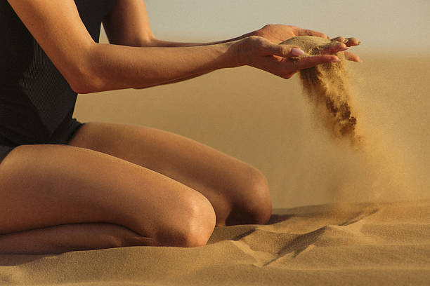 A serene moment captured as a woman kneels on golden desert sand, letting it effortlessly flow through her hands, depicting the passage of time and tranquility in nature. The warm light and soft textures illustrate a connection with the environment and evoke a sense of mindfulness and reflection.