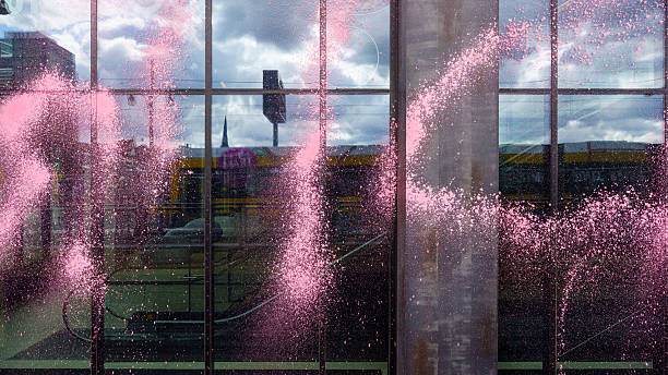 A glass building facade is splattered with bright pink paint, partially obstructing the reflection of the urban surroundings