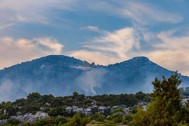 A Croatia Air Force Canadair CL-415 firefighting aircraft performs a water drop over a wildfire raging through rugged Dalmatian mountains. The dramatic scene captures thick smoke, pine forest, rocky terrain, and the precision timing of aerial firefighting. This powerful editorial image represents the growing challenge of wildfires in Southern Europe and the urgent need for climate resilience.
