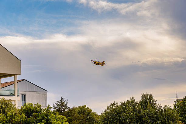 A Canadair firefighting plane flies low over a residential neighborhood in coastal Croatia during wildfire season. The contrast between modern buildings and the yellow aircraft highlights the urgency of fire prevention in populated areas. Ideal for editorial articles on climate response, emergency air support, or environmental issues in southern Europe.