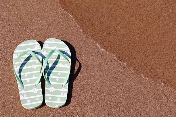 Close-up of a mature woman’s hand holding green flip flops while standing on the shore in a bikini. The sea is in the background with gentle waves and sparkling water. Concept of summer vacation, seaside relaxation, beach fashion, and healthy lifestyle.
