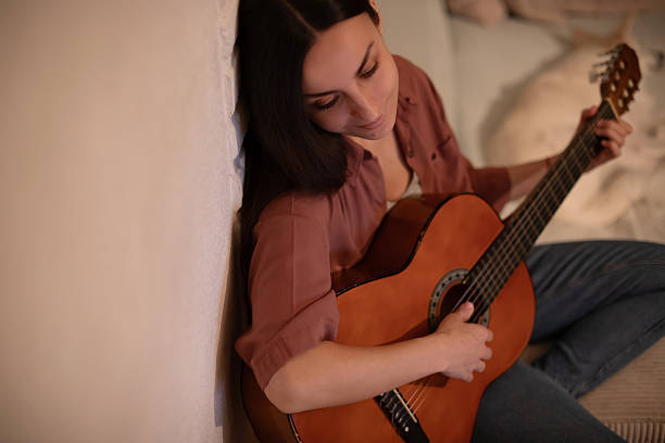 istock A young woman playing an acoustic guitar while sitting on a sofa in a cozy indoor setting. She is enjoying her time, immersed in music, with a relaxed expression and soft lighting surrounding her. 2220794820