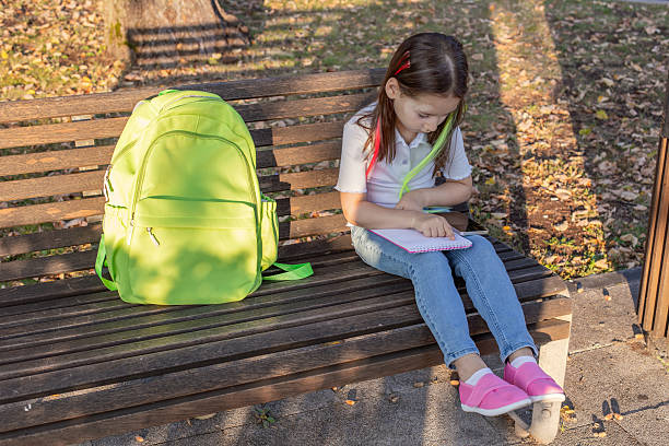 istock Young girl sitting on a park bench writing in notebook with green backpack 2220797986