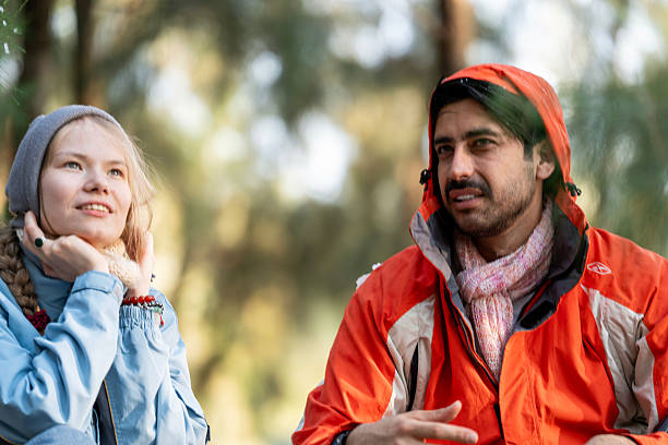 istock Happy Couple Playing with Snowfall in Winter Forest – Man Gently Touching Woman’s Hood While Both Laugh and Enjoy the Moment, Natural Light Scene with Snowflakes and Adventure Gear 2220817507
