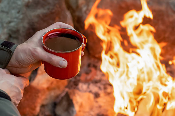 Camping lifestyle concept. Hands holding red enamel mug near a campfire. Hiking