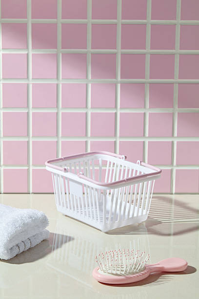 A transparent plastic basket with towel and bath sponge is placed on a glossy beige counter, set against a soft pink mosaic tile wall. A clean, minimal bathroom scene for wellness and home care themes.