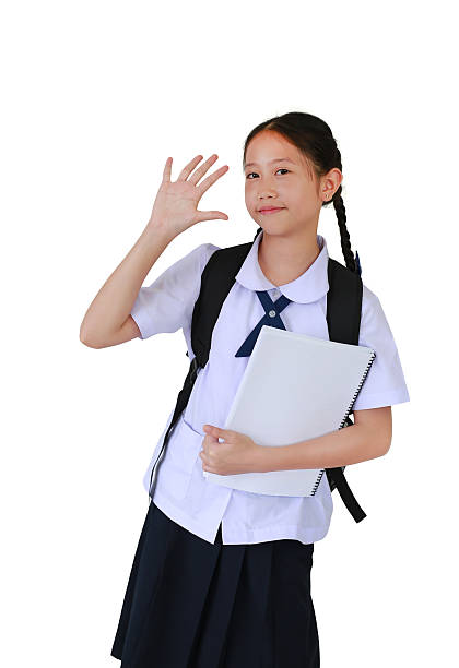 Smiling Asian student girl in school uniform waving hand hello while carrying backpack bag and holding a book against white background.