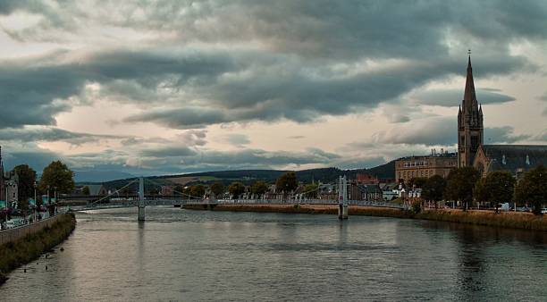 A picturesque view of Inverness, the capital of the Scottish Highlands, featuring the tranquil River Ness flowing through the city with the iconic Old High Church's spire rising prominently on the riverbank. The cloudy sky adds to the dramatic atmosphere of the Scottish landscape.