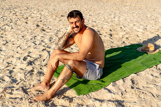 Young tattooed man sitting on a vibrant green towel, soaking up the sun and enjoying a relaxing day at the beach