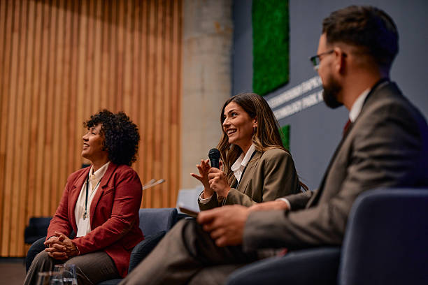 Businesswoman sitting at a conference. Speaking into a microphone while colleagues attentively listening to her insightful presentation. Fostering communication and collaboration among professionals