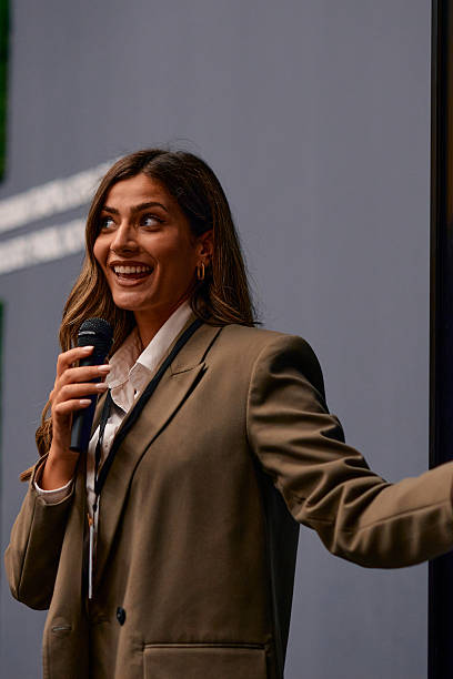 Businesswoman smiling and engaging with the audience at a conference, holding a microphone while dressed in a stylish brown suit, radiating professionalism and confidence