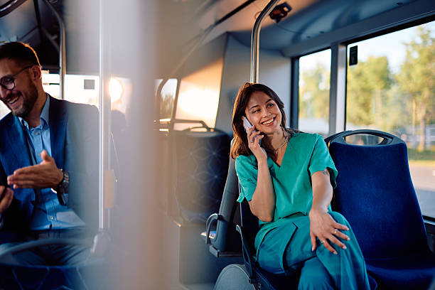 Young female nurse in scrubs enjoys her commute, talking on her smartphone while sitting on a bus, a businessman sits in the foreground, using his own phone