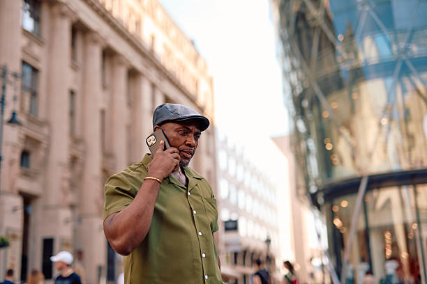 Stylish mature African American man wearing a green shirt and a leather cap is walking down a busy city street while talking on his smartphone