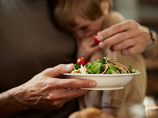 Close up of unrecognizable single father trying to feed his small boy with fresh salad while he is refusing to eat.
