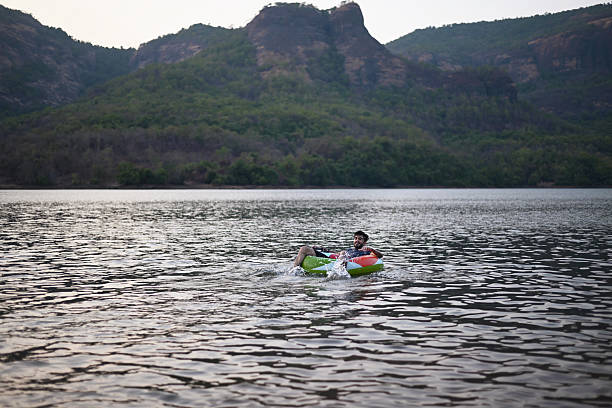 A young man lies back on an inflatable swim ring in a calm lake. The water reflects the soft evening light, with a rocky hills in the background.