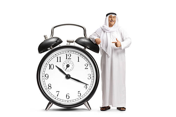 Middle eastern man in traditional clothes leaning on a big alarm clock and pointing isolated on white background