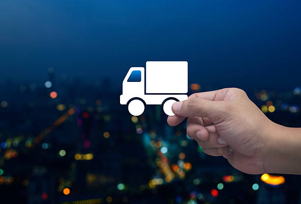 Hand holding delivery truck flat icon in fingers over blur colorful night light modern city tower and skyscraper, Business transportation service concept