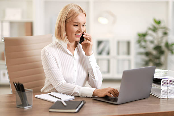 istock Smiling middle aged woman talking by smartphone at table in office 2220871510