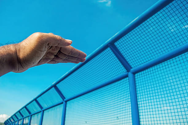 A human hand reaches outward, seemingly interacting with a long, vibrant blue mesh fence under a clear sky. Conveys themes of reaching, boundaries, and the human desire for connection or freedom