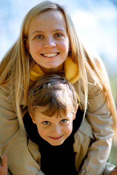 Blonde woman with long hair embraces her cheerful young son from behind on a bright day outdoors, both smiling warmly into the camera. Perfect for your advertisement and commercial using.