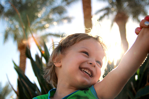 Cheerful toddler laughing under bright sunlight, surrounded by palm trees in a tropical outdoor setting.