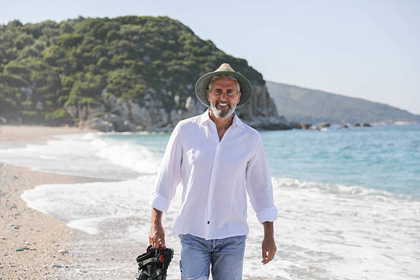 Mature Man Walking Barefoot on the Beach Holding Sandals