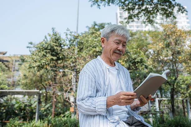 Senior man sits calmly on a park bench, fully absorbed in his book. Surrounded by nature, he enjoys a quiet moment of relaxation and mindfulness.