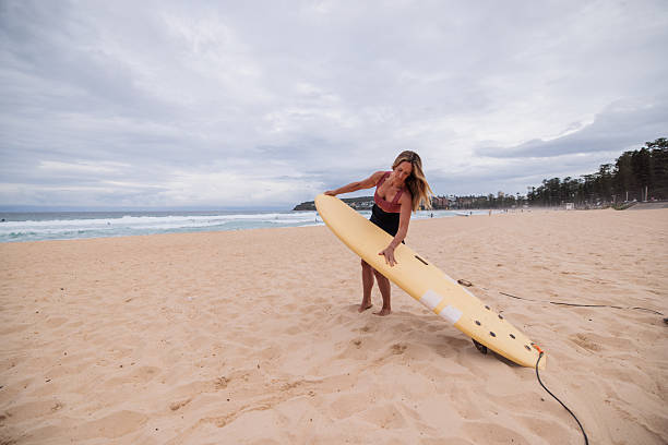 Woman with beach-ready gear prepares her board at the edge of the surf, getting ready for a ride on the waves at Manly Beach.