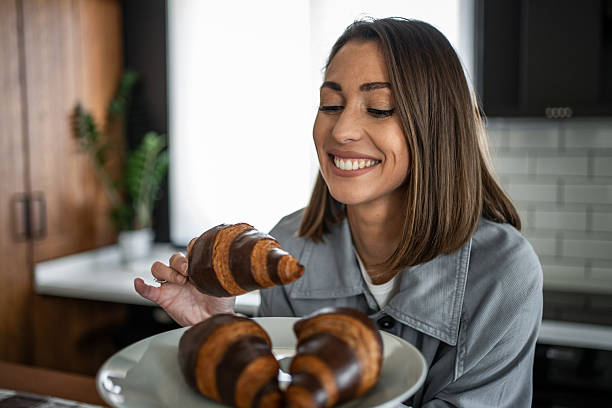 Cheerful young adult woman looking and smiling at chocolate croissant she is holding in her hand, while other chocolate croissants are on a plate in the kitchen