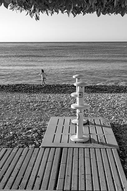 Altea. Alicante. Spain. Black and white photograph capturing an evocative summer beach vacation scene, with beach showers prominently in the foreground. Set on a pebble beach, the calm atmosphere is completed by the soft silhouette of a young girl playing out of focus near the waterline, evoking a quiet moment of coastal life.