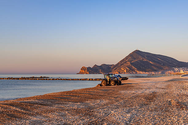 Altea. Alicante. Spain. Tractor cleaning the beach at dawn, revealing smooth sand and pebbles while a majestic mountain rises in the background, complemented by a breakwater stretching into the sea