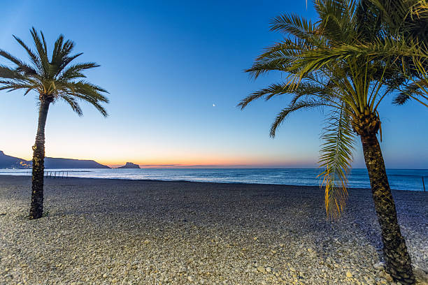 Altea. Alicante. Spain. Sunrise on the beach of Altea, Spain, with palm trees, pebbles, the silhouette of the Peñón de Ifach and the moon in the sky.