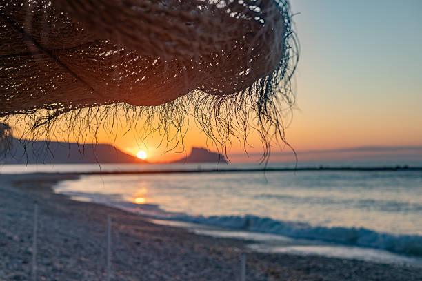 Altea. Alicante. Spain. Straw beach umbrella provides shade from the rising sun over a calm ocean, creating a serene and relaxing atmosphere