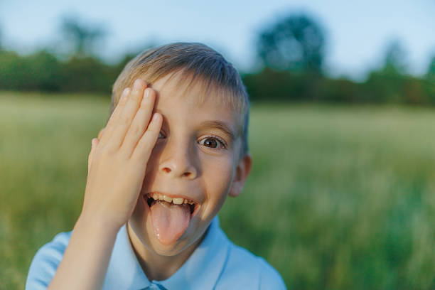 A cheerful little boy outdoors makes a silly face, tilting his head and sticking out his tongue. Playful summer energy with natural background and warm light.