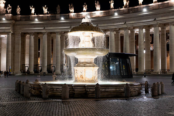 A nighttime view of the illuminated fountain in St. Peter’s Square, Vatican City, with cascading water and colonnades in the background