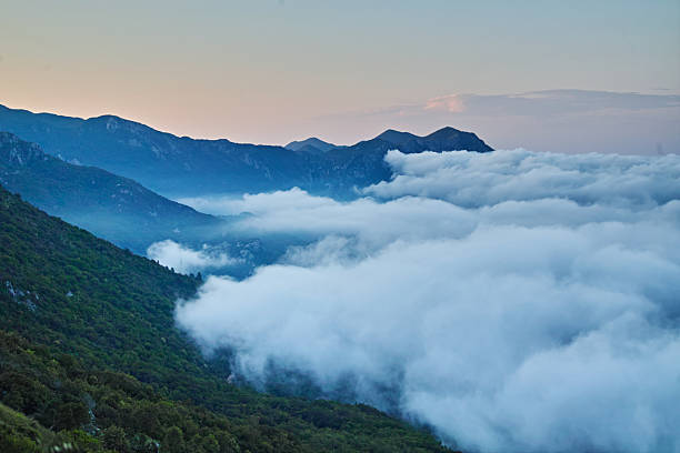 A breathtaking landscape view of majestic mountains partially shrouded in thick, low-lying clouds or fog, creating a mystical and serene atmosphere. The slopes are covered in lush greenery, and the sky hints at dusk or dawn colors.