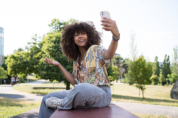 Smiling young woman with curly hair enjoying a video call on her smartphone while sitting on a bench in a sunny park surrounded by lush green trees and plants