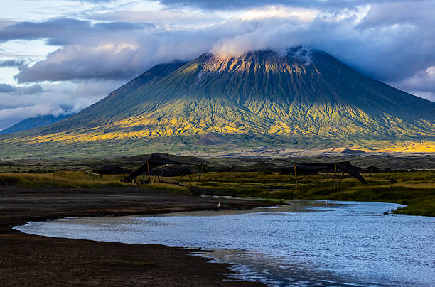 View of Ol Doinyo Lengai, an active stratovolcano in northern Tanzania, partially shrouded by clouds as streaks of sunlight illuminate its rugged slopes. In the foreground, a small stream winds through the arid landscape.