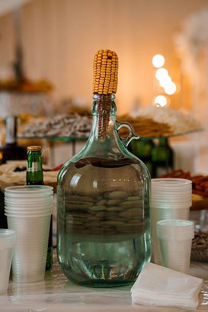 An atmospheric image showing a green glass jug adorned with corn atop a table alongside white cups and napkins, creating a cozy fall celebration ambiance