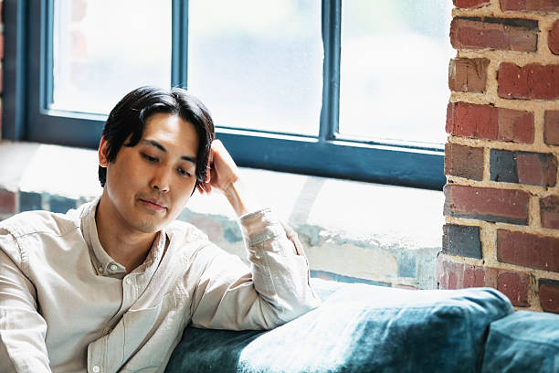 A mid adult Asian man in a coworking office space, sitting on a couch by a window, working on his laptop.