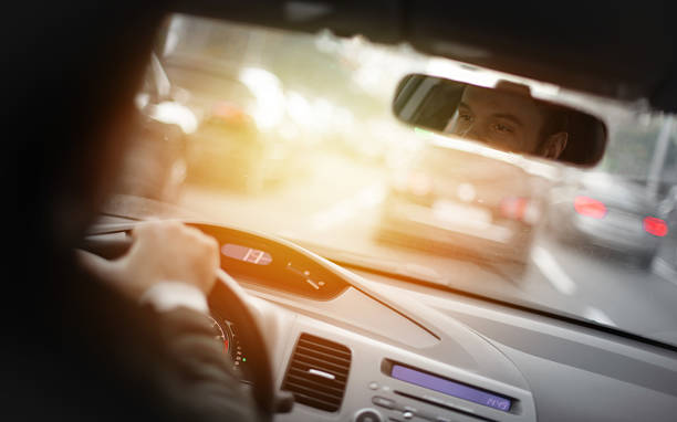 istock Man driving a car during daylight, visible in the rearview mirror with a focused expression, steering through traffic on a city road, with sunlight creating a warm, cinematic effect. 2221000987