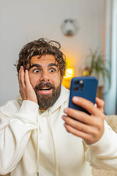 istock Surprised Indian bearded man using cellphone winning lottery sitting on sofa couch at home apartment 2221001310