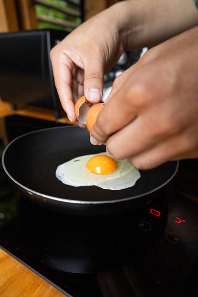 istock Hands holding cracked eggshell over frying egg in black pan, capturing the moment the egg meets the hot surface 2221001936